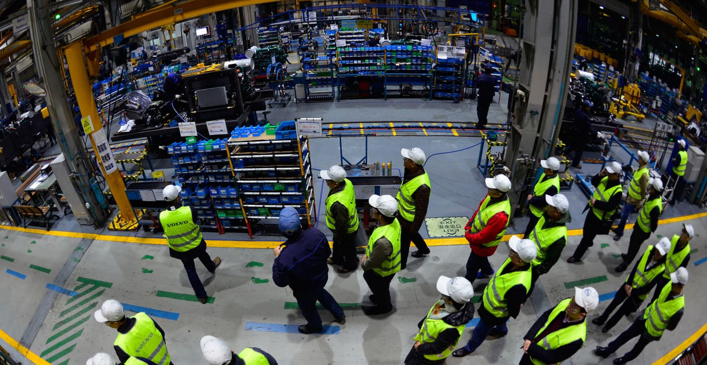 Visitors in hard hats and safety vests walking the floor of a large factory during an escorted plant tour.